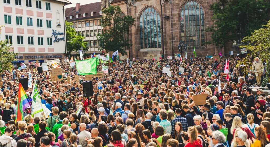 Eine große Menschenmenge, die an einer Demonstration oder Kundgebung gegen einen Streik teilnimmt. Die Veranstaltung findet in einer städtischen Umgebung statt, wie an den Gebäuden und der Architektur im Hintergrund zu erkennen ist. Eine der auffälligen Gebäude hat das Schild “Küchen Bosch” und eine Zeichnung einer Vase, was darauf hindeutet, dass sich die Kundgebung möglicherweise in einer deutschen Stadt befindet.
