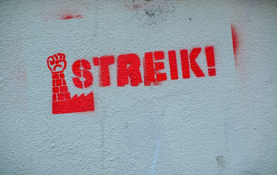 Ein Wandgraffiti mit dem Wort “STREIK!” in roter Farbe. Links neben dem Schriftzug ist ein Symbol zu sehen: eine rote Faust, die nach oben gereckt ist und deren Arm aus Ziegeln besteht. Der Arm ragt aus einem stilisierten Fabrikgebäude empor. Dieses Bild vermittelt eine klare Botschaft des Protests und der Arbeitersolidarität. Der Schriftzug und das Symbol sind beide in einem kräftigen, auffälligen Rot gehalten, was die Dringlichkeit und den Nachdruck des Themas eines Streik unterstreicht.