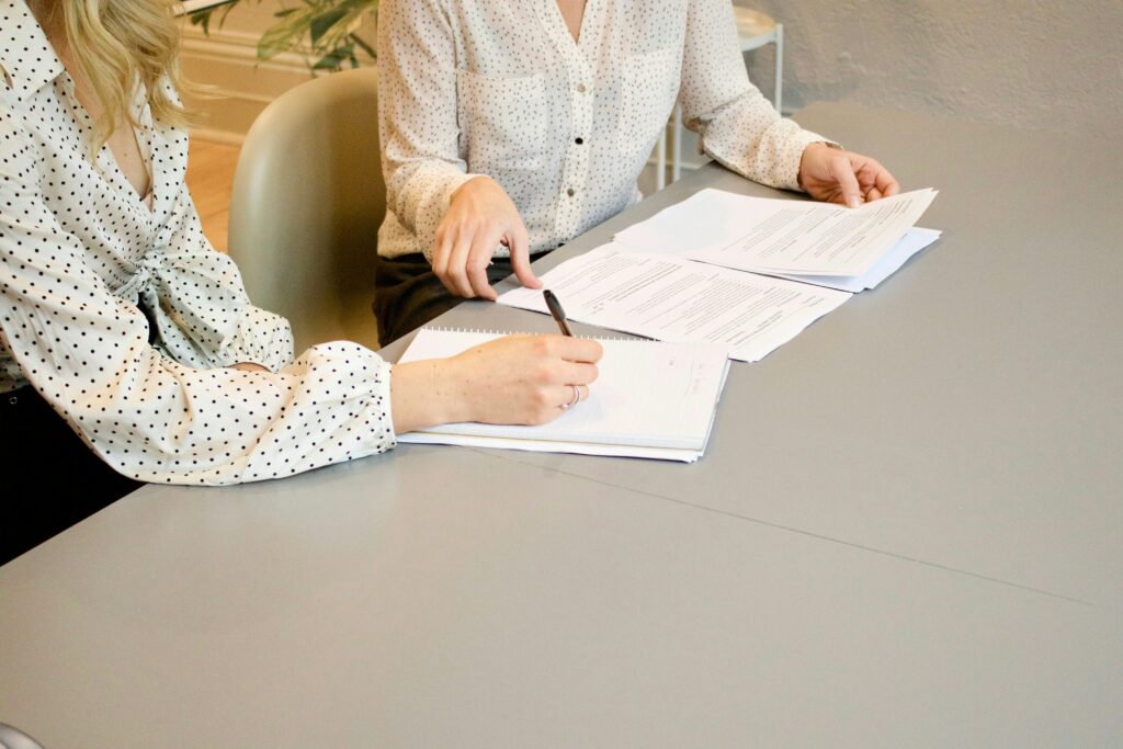 Das Bild zeigt zwei Personen, die an einem Tisch sitzen und über Dokumente und Notizen arbeiten. Beide tragen gepunktete Blusen und scheinen sich auf den Inhalt der Papiere zu konzentrieren, während eine Person mit einem Stift etwas notiert. Dieses Bild kann mit der Arbeitslosenversicherung in Verbindung gebracht werden, da es die sorgfältige Prüfung und Dokumentation von Unterlagen symbolisiert, die notwendig sind, um Ansprüche geltend zu machen und Leistungen der Arbeitslosenversicherung zu erhalten.