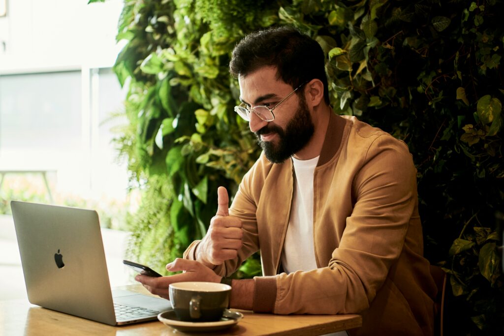 Ein Mann mit Brille und Bart, der in einem modernen, grünen Büro an einem Laptop arbeitet und mit einem Daumen hochgestikuliert, während er auf sein Smartphone schaut. Die Umgebung wirkt freundlich und einladend, mit einer Tasse Kaffee auf dem Tisch und üppigem Grün im Hintergrund. Dieses Bild wird mit dem Begriff Berufsgenossenschaft in Verbindung gebracht werden, da es die positive Arbeitsatmosphäre und die Unterstützung symbolisiert, die durch die Sicherheits- und Gesundheitsmaßnahmen der Berufsgenossenschaften gefördert werden, um ein angenehmes und sicheres Arbeitsumfeld zu gewährleisten.