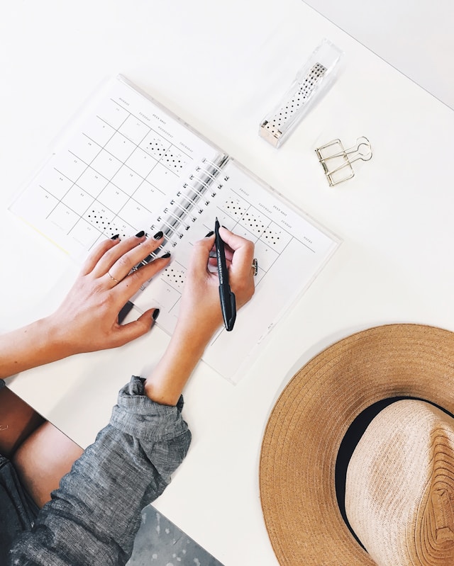 Eine Person, die einen Kalender oder Planer auf einem Schreibtisch benutzt und mit einem Stift Termine oder Aufgaben einträgt. Neben dem Planer liegen Büromaterialien wie eine Büroklammer, ein dekoratives Band und ein Strohhut, was auf eine bevorstehende Reise hinweist. Dieses Bild symbolisiert die sorgfältige Planung und Organisation, die für eine erfolgreiche Dienstreise notwendig ist, um alle beruflichen Verpflichtungen und Reisevorbereitungen effizient zu koordinieren.