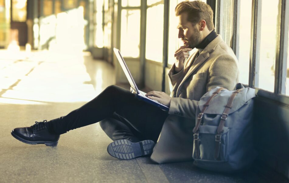 Ein Mann, der in einem gut beleuchteten, modernen Wartebereich auf dem Boden sitzt und konzentriert an einem Laptop arbeitet. Neben ihm liegt ein Rucksack, was darauf hinweist, dass er sich möglicherweise auf einer Reise befindet. Dieses Bild symbolisiert die typischen Situationen auf einer Dienstreise, bei der berufliche Aufgaben oft unterwegs erledigt werden müssen, während man auf den nächsten Flug oder Zug wartet.