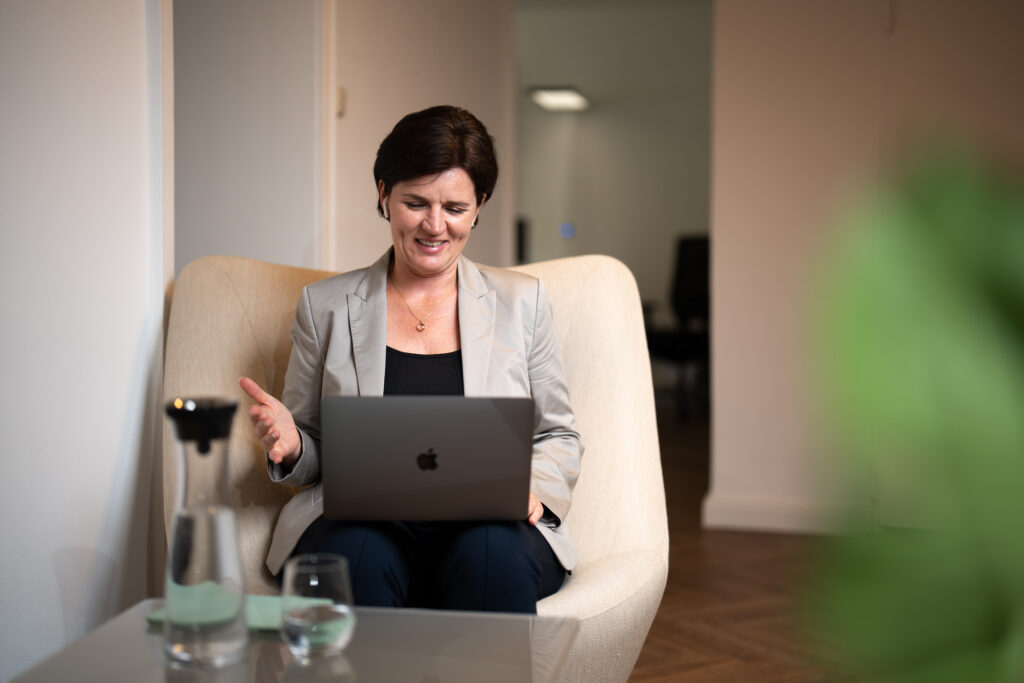 Das Bild zeigt eine Frau, die in einem Büro auf einem Stuhl sitzt und lächelnd an einem Laptop arbeitet. Neben ihr steht ein Tisch mit einer Wasserflasche und einem Glas. Die elektronische Arbeitsunfähigkeitsbescheinigung (eAU) erleichtert die Bearbeitung von Krankmeldungen, während moderne Lohnabrechnungssoftware sicherstellt, dass alle relevanten Daten schnell und effizient in die Lohnabrechnungen integriert werden können.