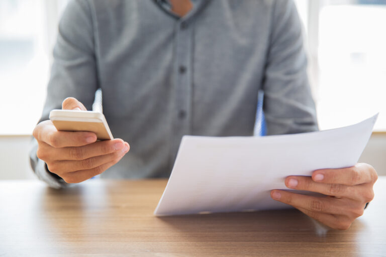 Eine Person, die ein Smartphone in der einen Hand und ein Dokument in der anderen Hand hält. Die Person scheint Informationen zu überprüfen oder zu bearbeiten. Dies lässt sich mit dem Thema Kurzarbeitergeld verbinden, da es zeigt, wie Arbeitnehmer und Arbeitgeber digitale Mittel nutzen können, um Anträge und erforderliche Unterlagen für die Beantragung und Verwaltung von Kurzarbeitergeld effizient zu bearbeiten.