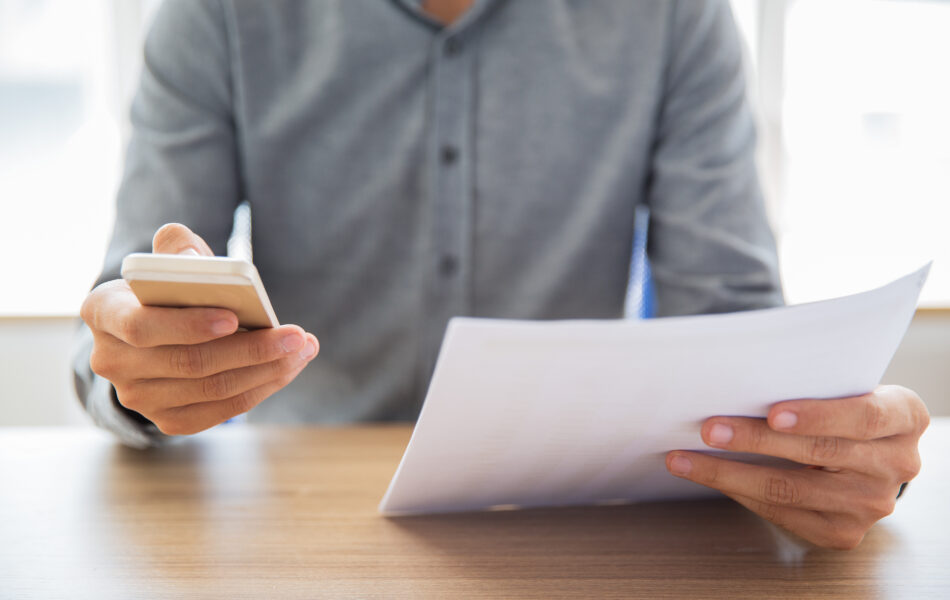 Eine Person, die ein Smartphone in der einen Hand und ein Dokument in der anderen Hand hält. Die Person scheint Informationen zu überprüfen oder zu bearbeiten. Dies lässt sich mit dem Thema Kurzarbeitergeld verbinden, da es zeigt, wie Arbeitnehmer und Arbeitgeber digitale Mittel nutzen können, um Anträge und erforderliche Unterlagen für die Beantragung und Verwaltung von Kurzarbeitergeld effizient zu bearbeiten.