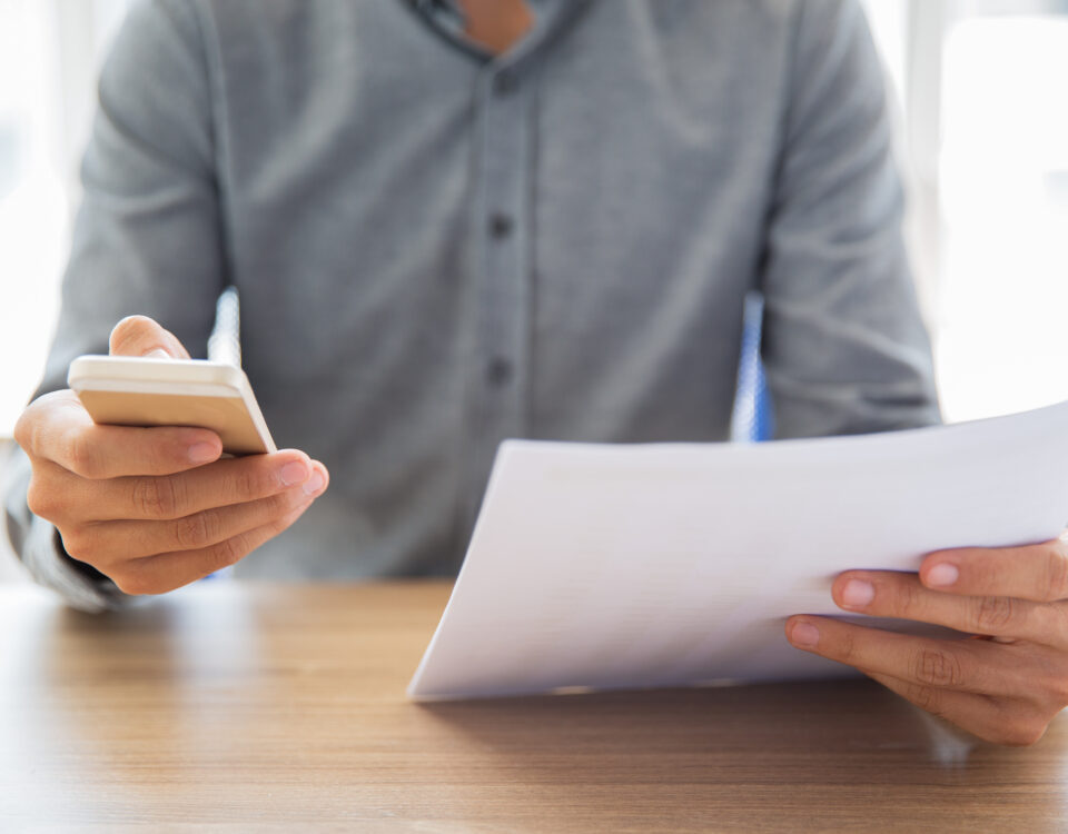 Eine Person, die ein Smartphone in der einen Hand und ein Dokument in der anderen Hand hält. Die Person scheint Informationen zu überprüfen oder zu bearbeiten. Dies lässt sich mit dem Thema Kurzarbeitergeld verbinden, da es zeigt, wie Arbeitnehmer und Arbeitgeber digitale Mittel nutzen können, um Anträge und erforderliche Unterlagen für die Beantragung und Verwaltung von Kurzarbeitergeld effizient zu bearbeiten.