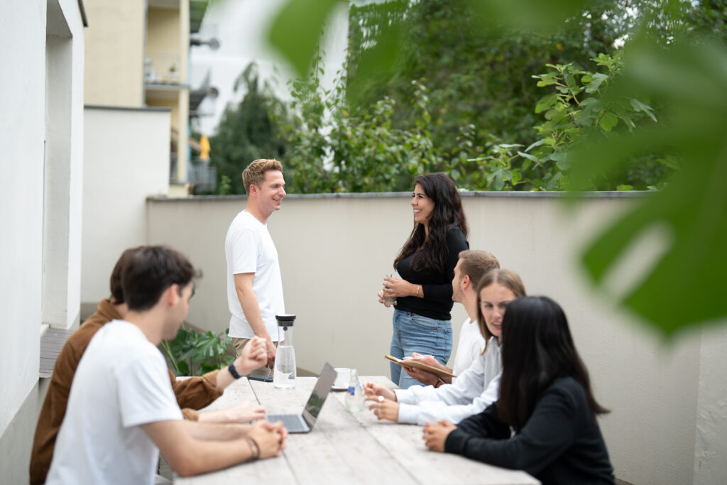 Eine Gruppe von sechs Personen sitzt und steht um einen Tisch in einer entspannten Outdoor-Umgebung, wobei einige diskutieren und Notizen machen. Die Integration vermögenswirksamer Leistungen in die Lohnabrechnungen erfordert präzise Berechnungen und Abstimmungen, um sicherzustellen, dass die zusätzlichen finanziellen Vorteile korrekt an die Mitarbeiter weitergegeben werden. Gespräche in solchen informellen Meetings können dazu beitragen, das Verständnis und die Umsetzung dieser Leistungen im Unternehmen zu verbessern.