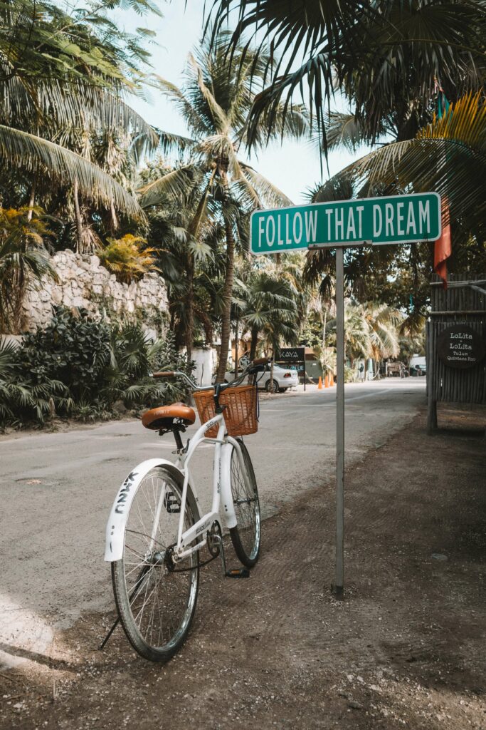 Das Bild zeigt ein weißes Fahrrad, das an einem Straßenschild mit der Aufschrift “FOLLOW THAT DREAM” inmitten einer tropischen Umgebung mit vielen Palmen angelehnt ist. Die Szene strahlt ein Gefühl von Abenteuer und Zielstrebigkeit aus, indem sie dazu ermutigt, seinen Träumen zu folgen. Der Verpflegungsmehraufwand kann als notwendiger Begleiter auf solchen Reisen gesehen werden, da er die zusätzlichen Kosten für Verpflegung abdeckt und es ermöglicht, sich voll auf das Erreichen der eigenen Ziele und Träume zu konzentrieren.