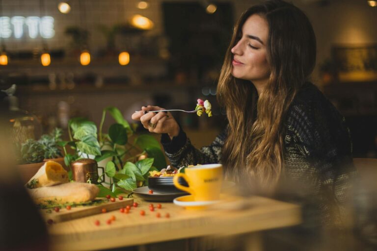 Eine junge Frau mit langen, welligen Haaren, die an einem gemütlichen Tisch in einem Café oder Restaurant sitzt. Sie lächelt entspannt und genießt offenbar ihre Mahlzeit, während sie einen Bissen Essen auf einer Gabel hält. Ihre Augen sind leicht geschlossen, was auf einen Moment des Genusses und der Zufriedenheit hindeutet.
