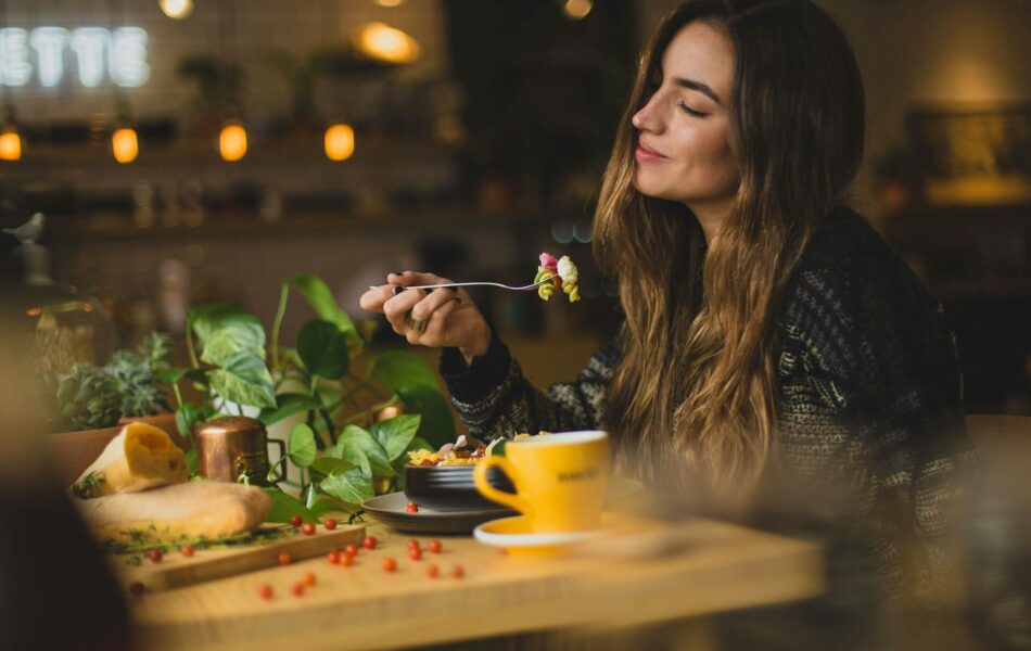 Eine junge Frau mit langen, welligen Haaren, die an einem gemütlichen Tisch in einem Café oder Restaurant sitzt. Sie lächelt entspannt und genießt offenbar ihre Mahlzeit, während sie einen Bissen Essen auf einer Gabel hält. Ihre Augen sind leicht geschlossen, was auf einen Moment des Genusses und der Zufriedenheit hindeutet.