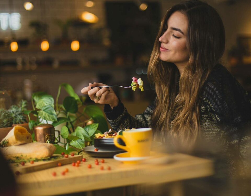 Eine junge Frau mit langen, welligen Haaren, die an einem gemütlichen Tisch in einem Café oder Restaurant sitzt. Sie lächelt entspannt und genießt offenbar ihre Mahlzeit, während sie einen Bissen Essen auf einer Gabel hält. Ihre Augen sind leicht geschlossen, was auf einen Moment des Genusses und der Zufriedenheit hindeutet.