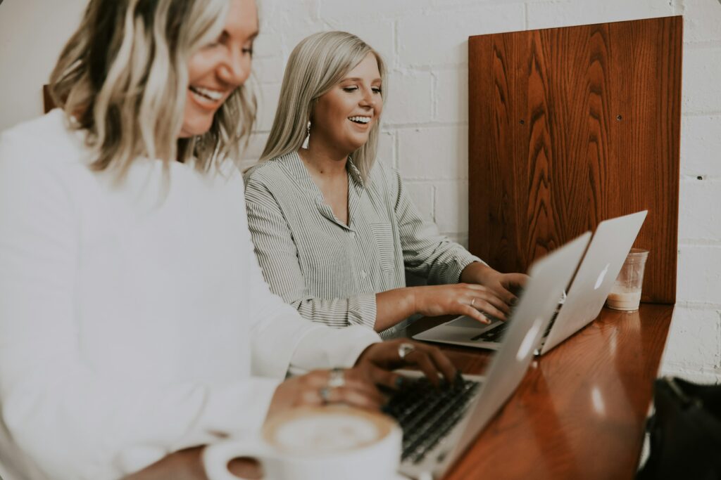 Das Bild zeigt zwei Frauen, die zusammenarbeiten und dabei lächeln, während sie an ihren Laptops sitzen. Die entspannte Atmosphäre und der offene Austausch wirken motivierend und unterstützend. In Verbindung mit “Mutterschutz” symbolisiert das Bild die Rückkehr an den Arbeitsplatz nach der Elternzeit, wobei ein flexibles und kollegiales Arbeitsumfeld die Vereinbarkeit von Beruf und Familie erleichtert.