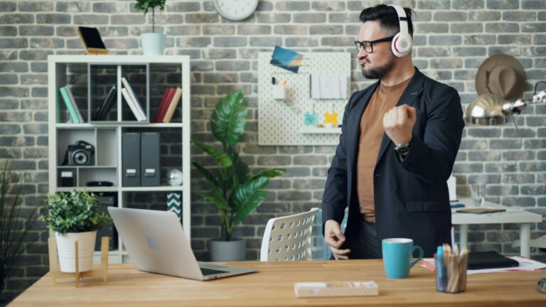 Ein Mann in legerem Business-Outfit steht in einem modernen Büro und bewegt sich energiegeladen zur Musik, die er über seine Kopfhörer hört. Der Schreibtisch vor ihm ist mit einem Laptop, einer Tasse und Büromaterialien ausgestattet, was eine entspannte und kreative Arbeitsatmosphäre vermittelt. Geldwerter Vorteil verweist hier auf mögliche Arbeitgeberleistungen, wie etwa die Bereitstellung eines modernen und komfortablen Arbeitsumfelds, das die Zufriedenheit und Motivation der Mitarbeitenden steigern kann.