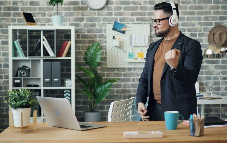 Ein Mann in legerem Business-Outfit steht in einem modernen Büro und bewegt sich energiegeladen zur Musik, die er über seine Kopfhörer hört. Der Schreibtisch vor ihm ist mit einem Laptop, einer Tasse und Büromaterialien ausgestattet, was eine entspannte und kreative Arbeitsatmosphäre vermittelt. Geldwerter Vorteil verweist hier auf mögliche Arbeitgeberleistungen, wie etwa die Bereitstellung eines modernen und komfortablen Arbeitsumfelds, das die Zufriedenheit und Motivation der Mitarbeitenden steigern kann.