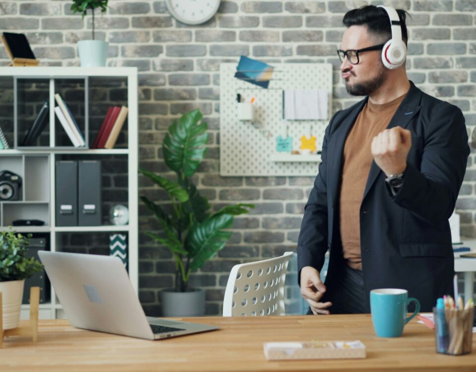 Ein Mann in legerem Business-Outfit steht in einem modernen Büro und bewegt sich energiegeladen zur Musik, die er über seine Kopfhörer hört. Der Schreibtisch vor ihm ist mit einem Laptop, einer Tasse und Büromaterialien ausgestattet, was eine entspannte und kreative Arbeitsatmosphäre vermittelt. Geldwerter Vorteil verweist hier auf mögliche Arbeitgeberleistungen, wie etwa die Bereitstellung eines modernen und komfortablen Arbeitsumfelds, das die Zufriedenheit und Motivation der Mitarbeitenden steigern kann.