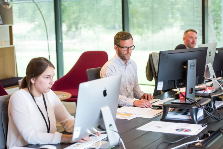 Drei Personen arbeiten konzentriert an ihren Computern in einem modernen Büro mit hellen Fenstern im Hintergrund. Der Fokus auf die Arbeitsumgebung deutet auf die administrative Verarbeitung von Daten hin. Steueridentifikationsnummer verweist hier auf die Notwendigkeit, eindeutige und korrekte Daten, wie die Steuer-ID der Mitarbeitenden, im System zu erfassen, um eine reibungslose Lohn- und Gehaltsabrechnung sicherzustellen.