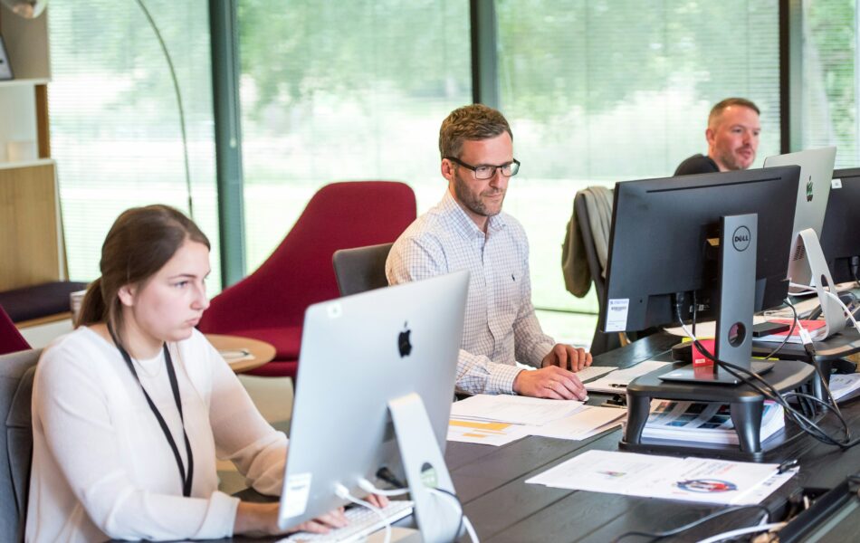 Drei Personen arbeiten konzentriert an ihren Computern in einem modernen Büro mit hellen Fenstern im Hintergrund. Der Fokus auf die Arbeitsumgebung deutet auf die administrative Verarbeitung von Daten hin. Steueridentifikationsnummer verweist hier auf die Notwendigkeit, eindeutige und korrekte Daten, wie die Steuer-ID der Mitarbeitenden, im System zu erfassen, um eine reibungslose Lohn- und Gehaltsabrechnung sicherzustellen.