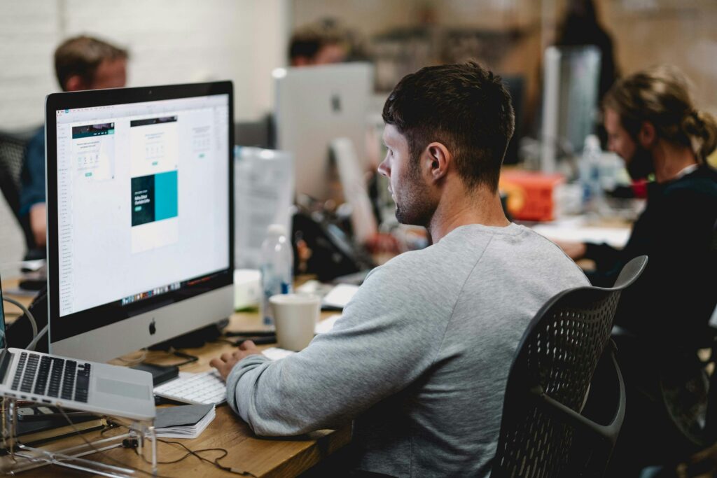 Ein Mitarbeiter sitzt konzentriert an einem Schreibtisch vor einem großen Monitor und arbeitet an digitalen Dokumenten. Im Hintergrund sind weitere Personen bei der Arbeit zu sehen. Sonderzahlungen verweisen hier auf die Aufgabe der Lohnbuchhaltung, solche Zusatzleistungen korrekt zu berechnen und zuverlässig in die monatlichen Gehaltsabrechnungen zu integrieren.