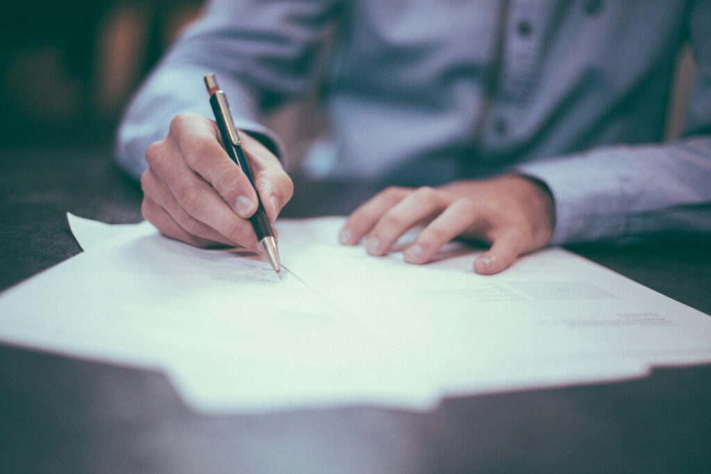 Eine Person in einem Hemd sitzt an einem Tisch und schreibt mit einem Stift auf ein Dokument, wobei mehrere Papiere vor ihr liegen. Die Szene vermittelt Professionalität und dokumentarische Genauigkeit. Sonderzahlungen verweisen hier auf die vertragliche oder gesetzliche Festlegung zusätzlicher Leistungen wie Urlaubs- oder Weihnachtsgeld, die schriftlich geregelt und dokumentiert werden müssen.