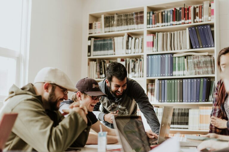 Eine Gruppe junger Menschen sitzt in einem hellen Raum an einem Tisch, arbeitet gemeinsam an Laptops und zeigt Freude und Begeisterung. Bücherregale im Hintergrund deuten auf einen Lern- oder Studienkontext hin. Studienbegleitetes Arbeiten verdeutlicht hier die Möglichkeit, Studium und praktische Teamarbeit zu kombinieren, um Wissen effektiv anzuwenden und wertvolle berufliche Erfahrungen zu sammeln.